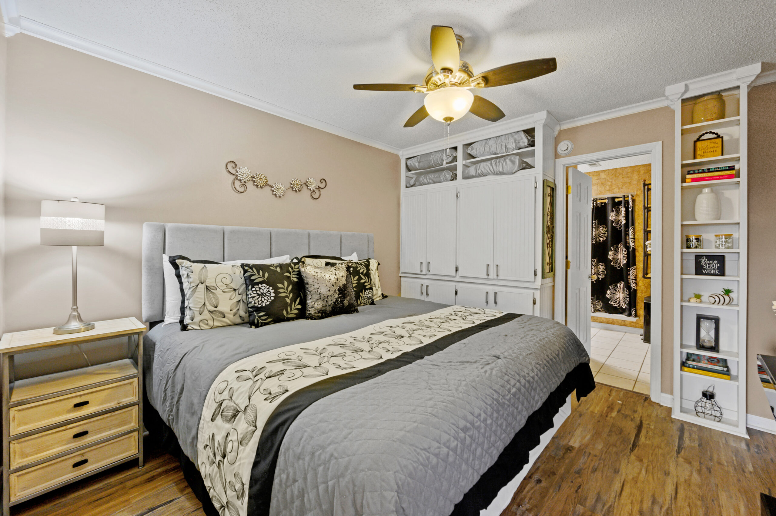 A king sized bed with grey, black, and white linens sits on a rustic wooden floor. Crisp, white built in bookshelves and armoire line the far wall. They are separated by a door to the ensuite full bath. A lighted ceiling fan is in the middle of the room, and a side table with lamp are to the left of the bed.