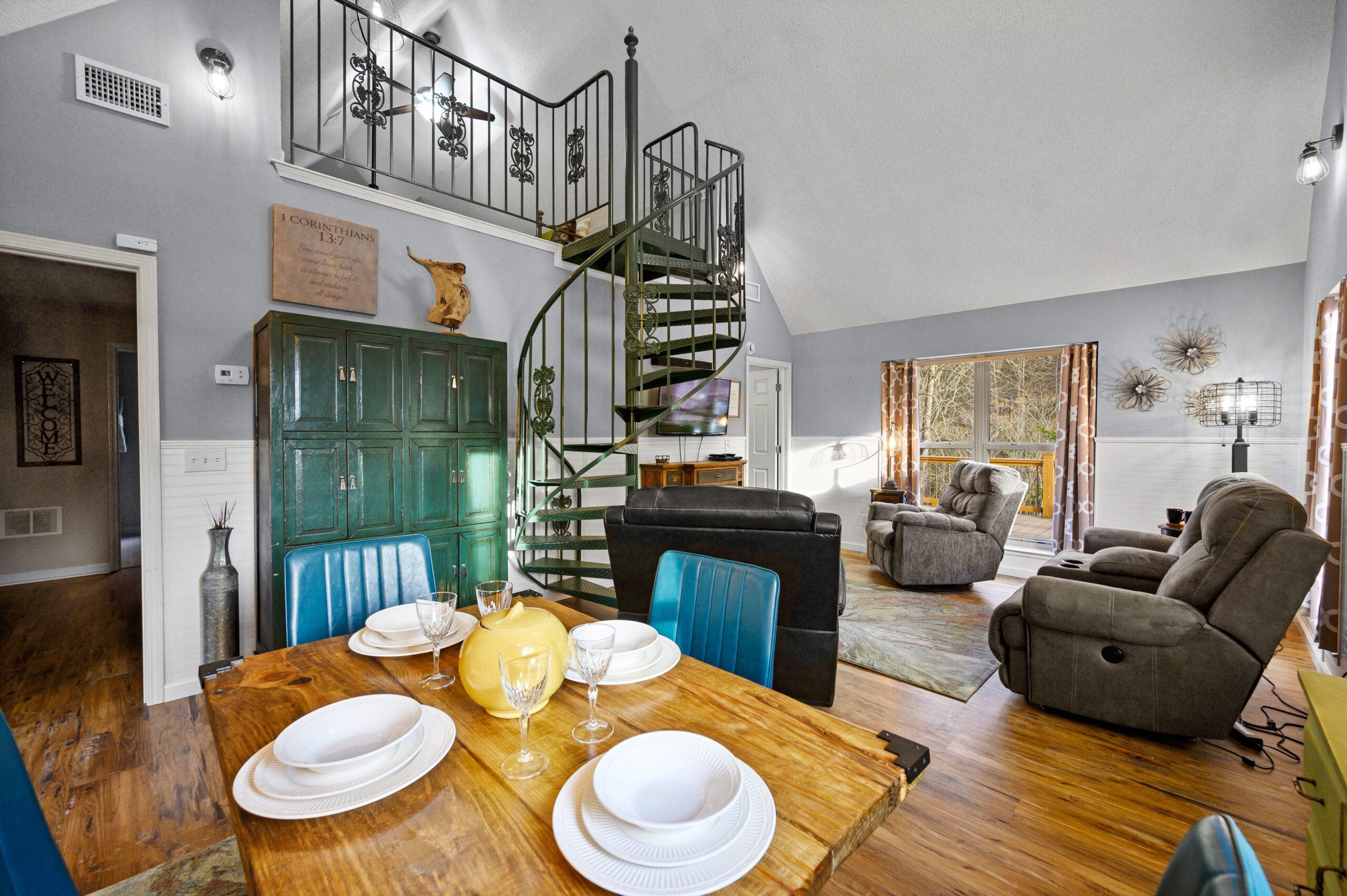 The main interior living space with industrial features. A wooden dining table with metal accents is set for 4, surrounded by teal mid-century dining chairs. Behind the custom made green circular staircase is the living area, with 4 oversized power recliners oriented to the TV.